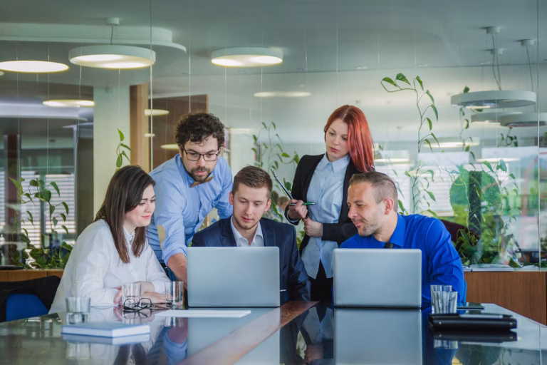 Group of professionals collaborating at a conference table with laptops, discussing ideas in a modern office setting, reflecting teamwork and productivity.